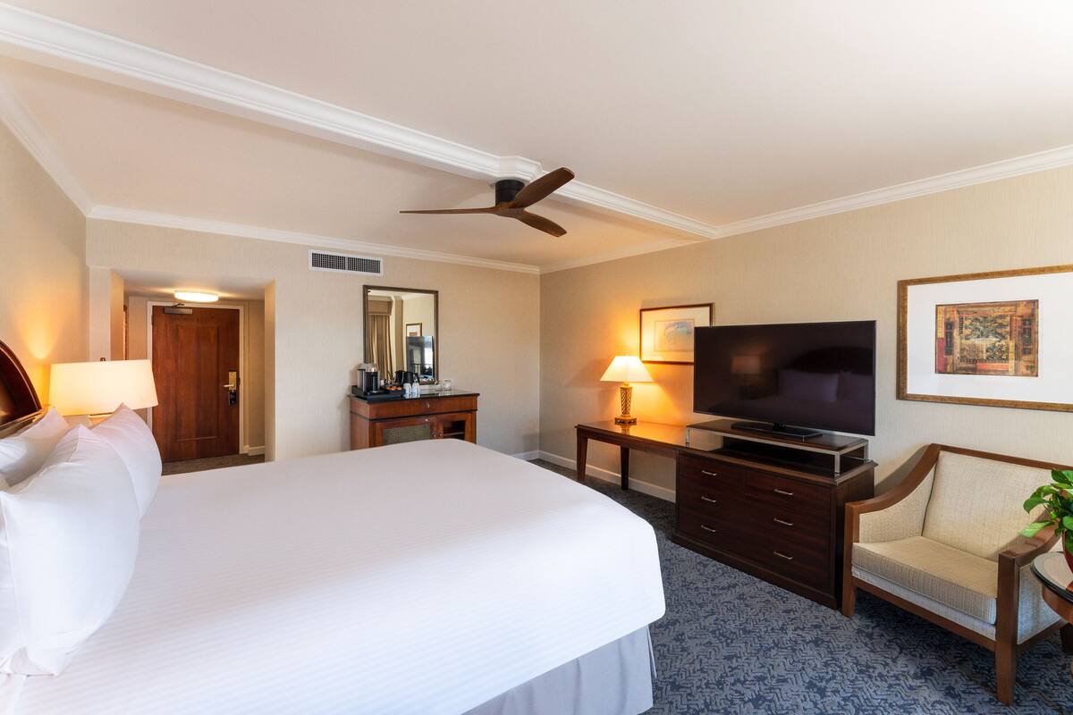 A hotel room with a large bed, a television, a desk with a lamp, a chair, and a ceiling fan.
