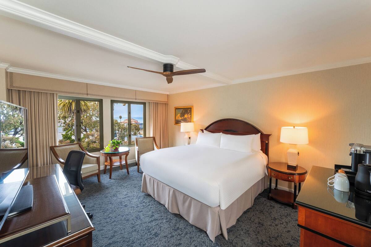A hotel room with a large bed, a desk, a chair, a small table with a plant, a coffee maker, and a lamp.