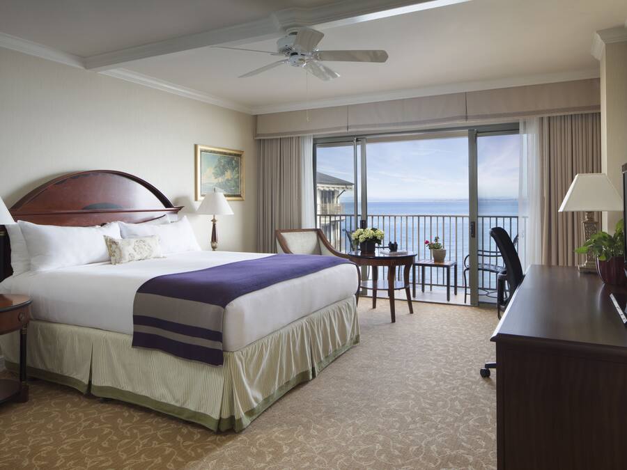A hotel room with a large bed, a ceiling fan, a TV, a desk, and a balcony with a view of the ocean.