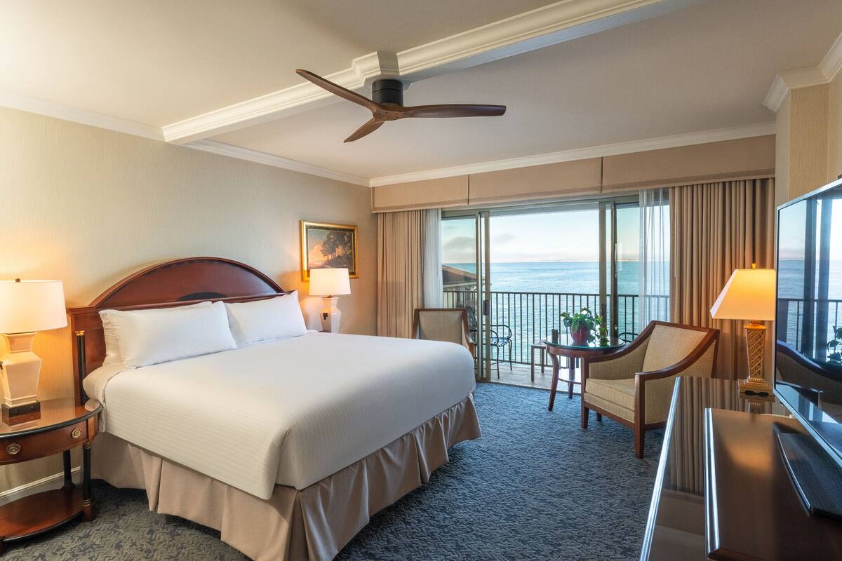 A hotel room with a large bed, two bedside tables with lamps, a desk with a chair, a TV, and a balcony with a view of the ocean.