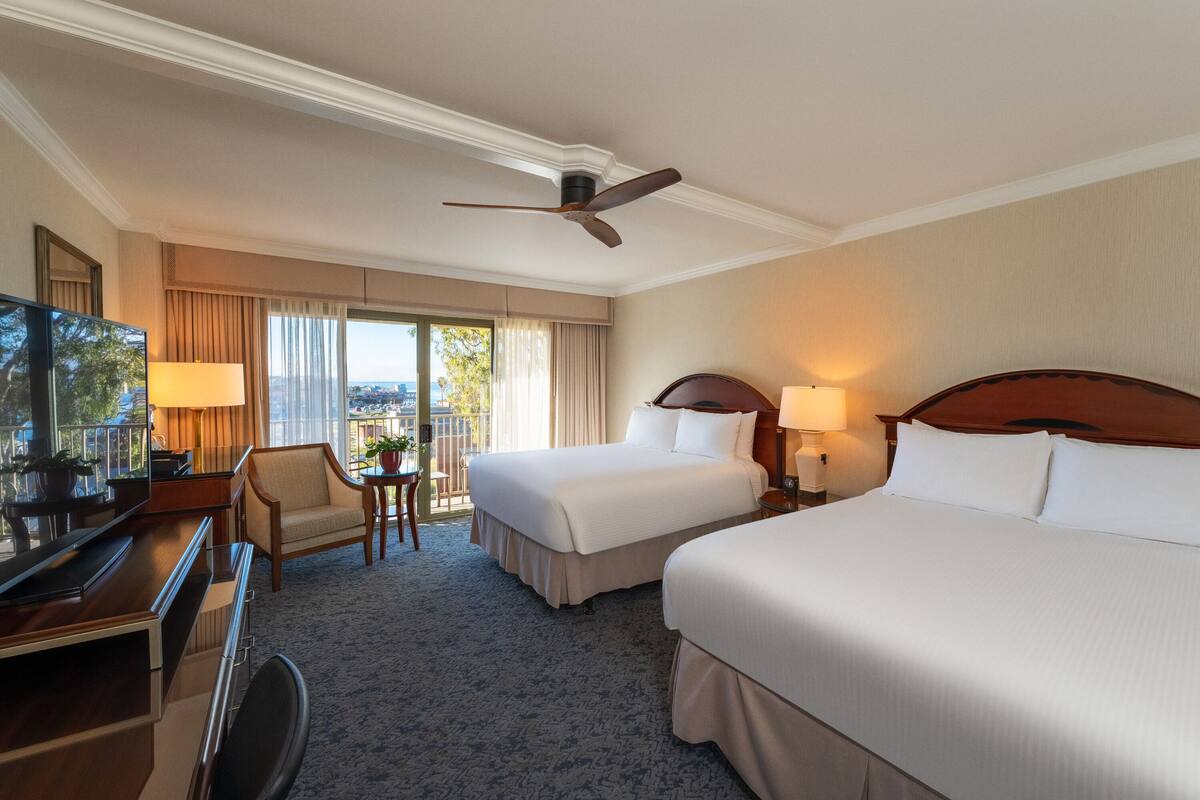 A hotel room with two beds, a desk with a chair, a TV, and a balcony with a view.