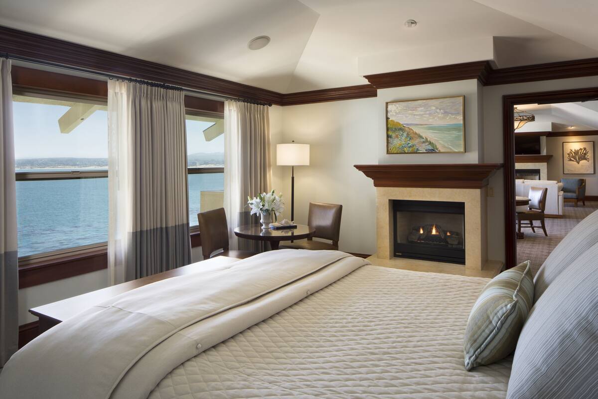 A hotel room with a large bed, a fireplace, a dining area, and a view of the sea.
