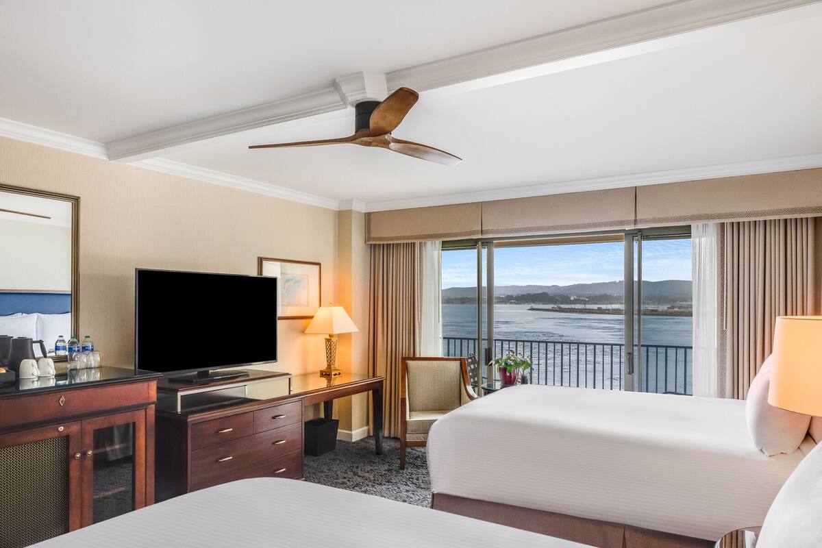 A hotel room with two beds, a TV, a desk with a lamp, and a view of the water.