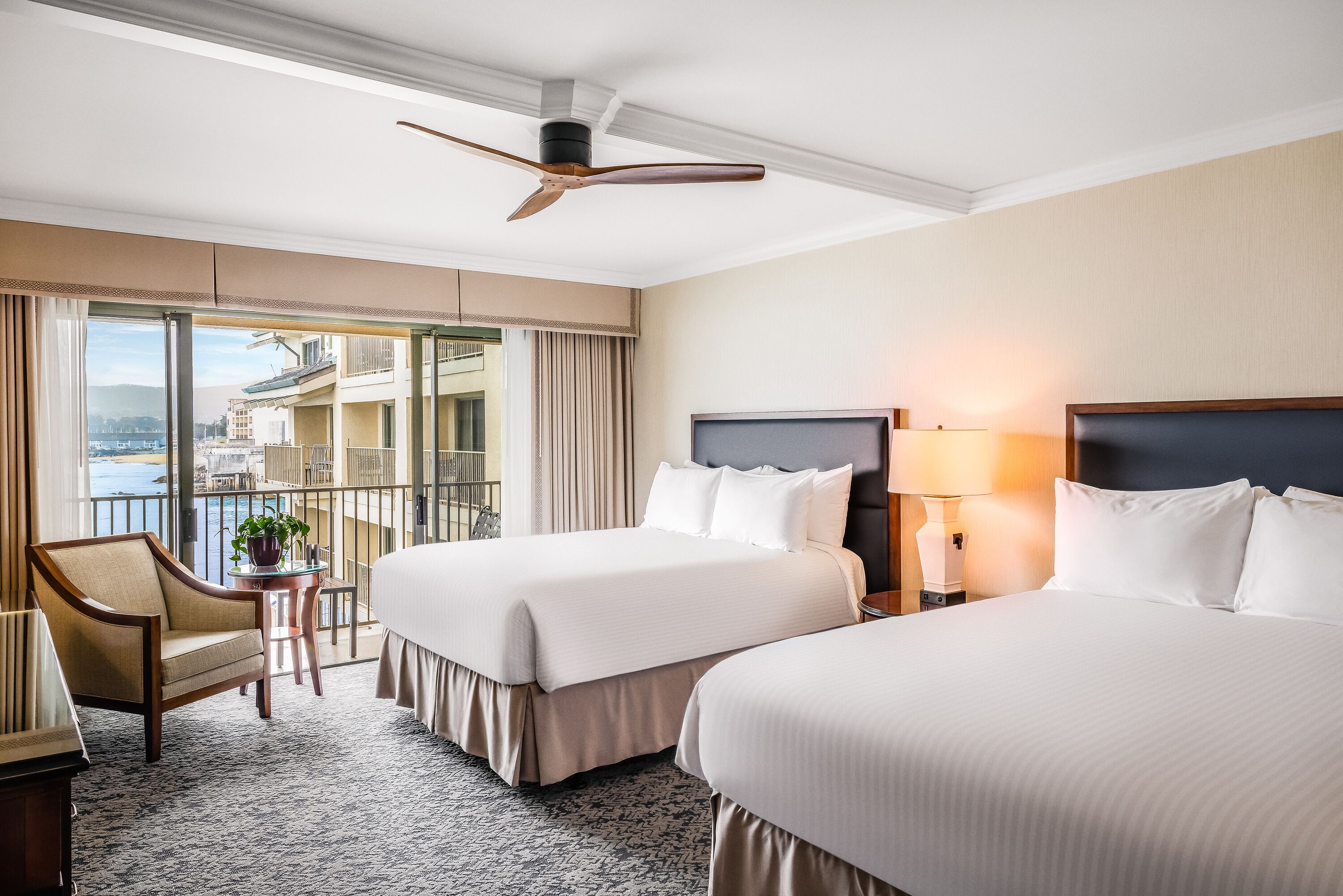 A hotel room with two beds, a chair, a small table with a plant, a balcony with a view of buildings and water, and a ceiling fan.