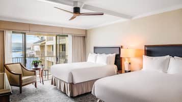 A hotel room with two beds, a chair, a small table with a plant, a balcony with a view of buildings and water, and a ceiling fan.