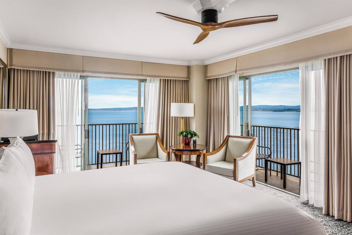 A hotel room with a large bed, two chairs, a small table with a plant, and a balcony with a view of the sea.