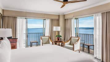 Quarto de hotel com uma cama grande, duas cadeiras, uma mesinha com uma planta e uma varanda com vista para o mar.