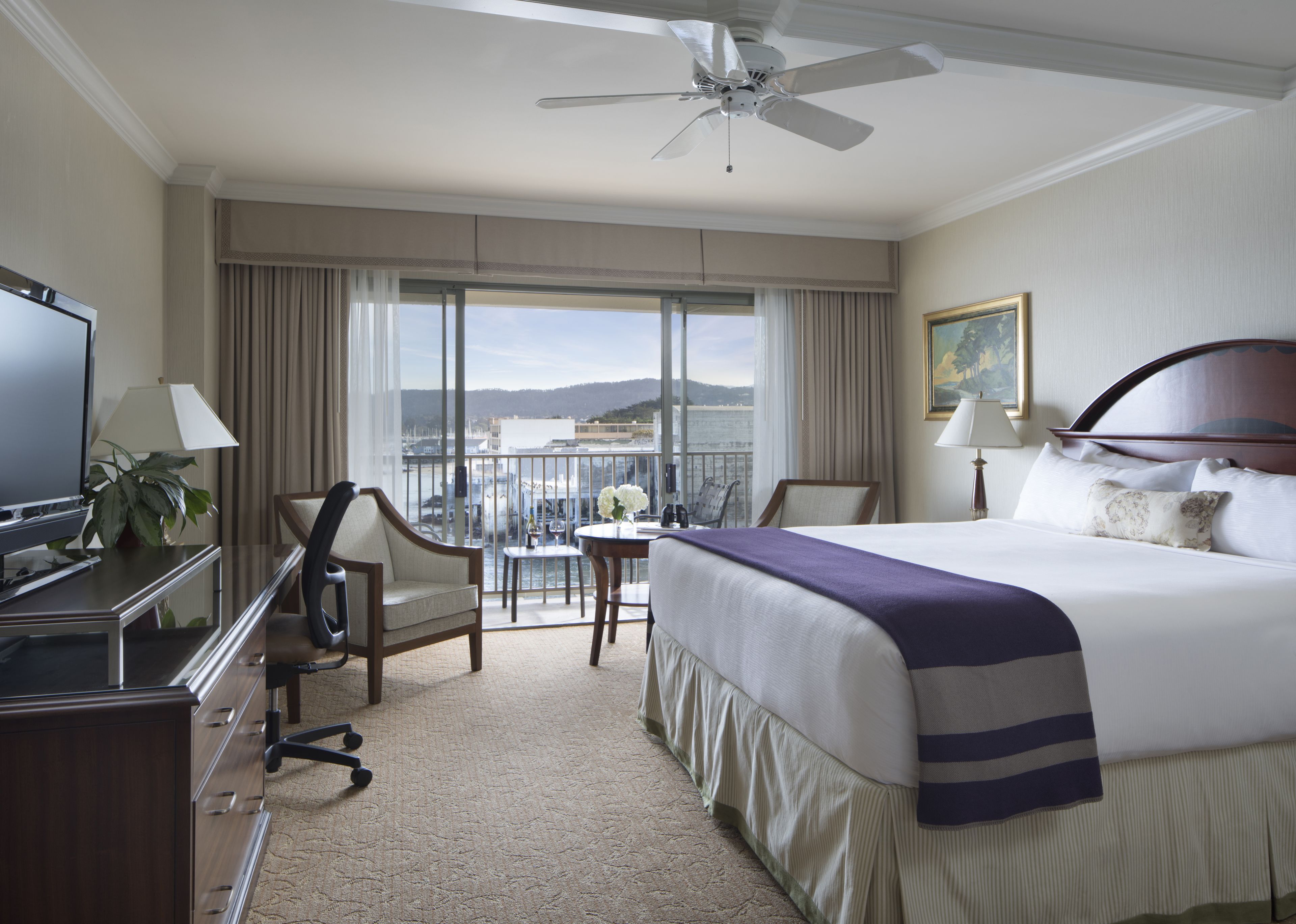 A hotel room with a large bed, a desk with a TV, a chair, a small table with a vase, and a balcony with a view of the water and hills.
