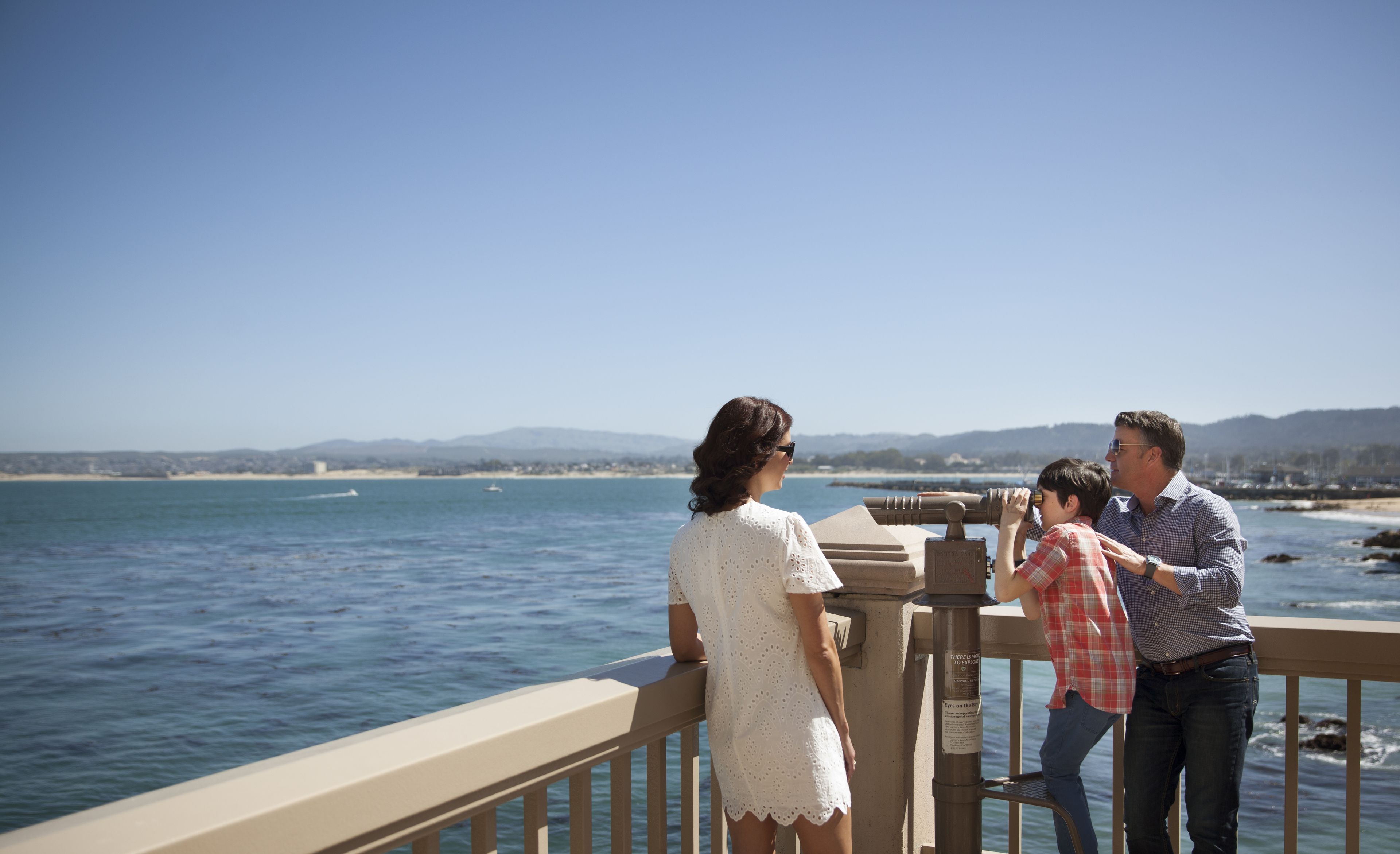 A couple and a child using binoculars on a balcony overlooking a coastal view.