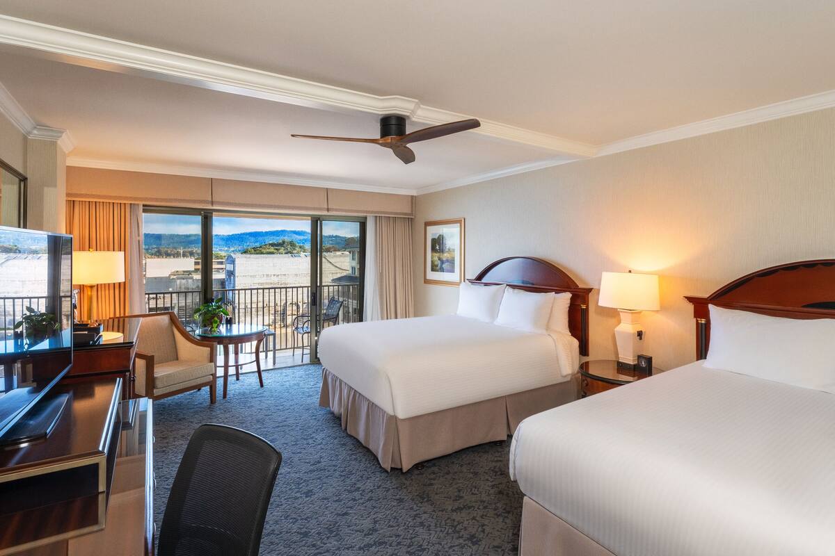 A hotel room with two beds, a desk, a chair, a balcony with a view, and a ceiling fan.