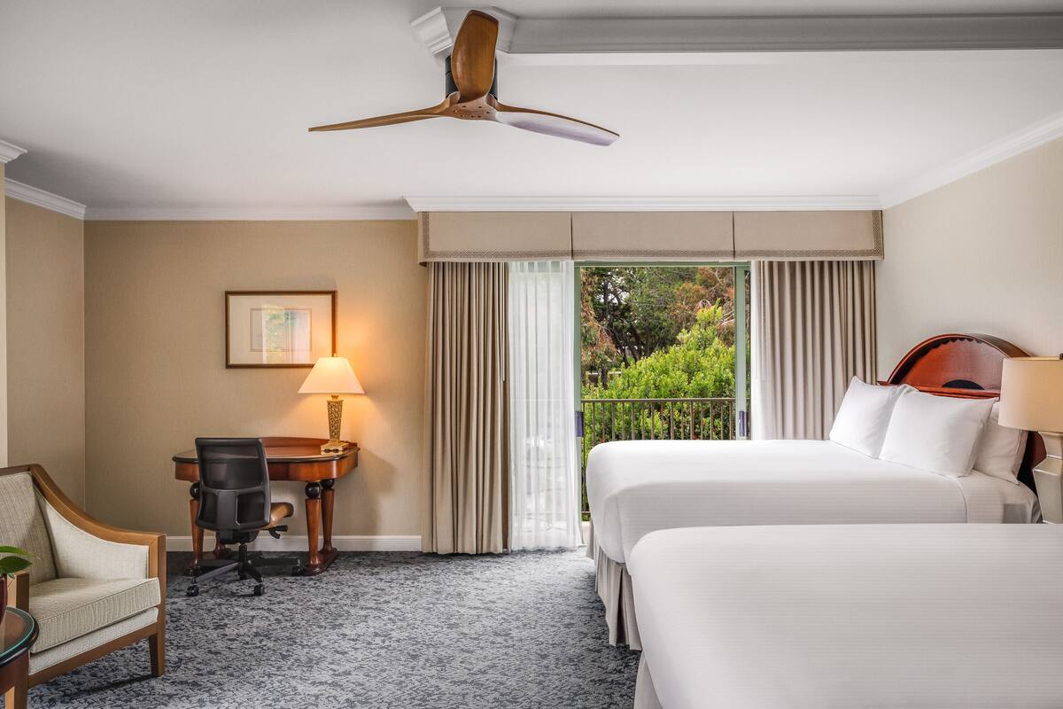 A hotel room with two beds, a desk with a chair, a ceiling fan, and a window with curtains.