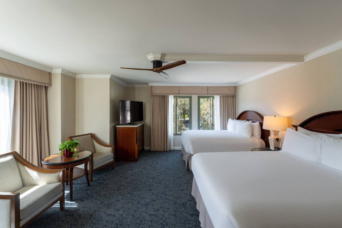 A hotel room with two beds, a TV, a desk with a plant, and a ceiling fan.