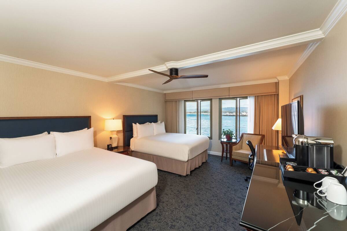 A hotel room with two beds, a desk with a coffee maker, a TV, and a view of the sea.