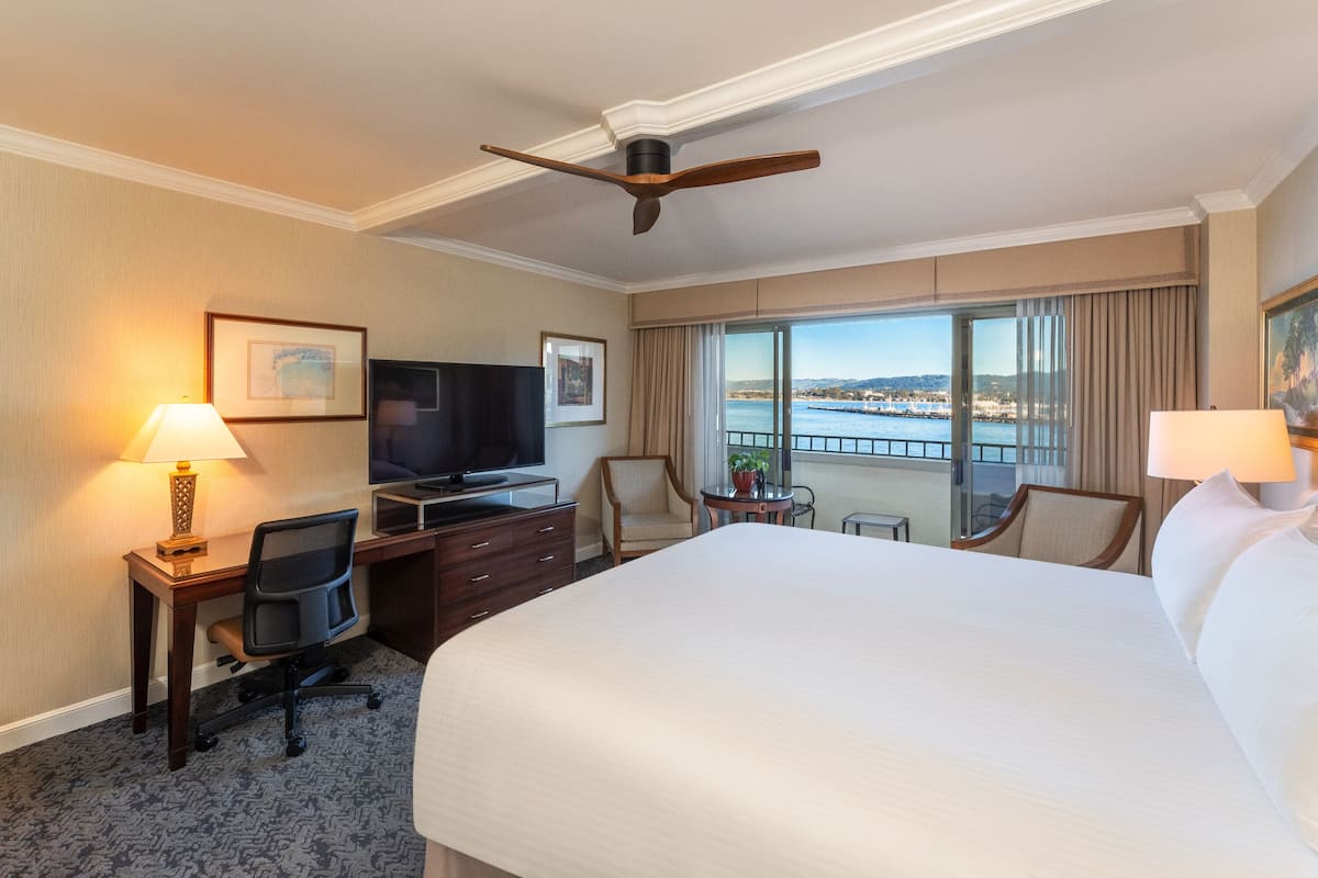 A hotel room with a large bed, a desk with a computer, a television, and a view of a body of water through large windows.