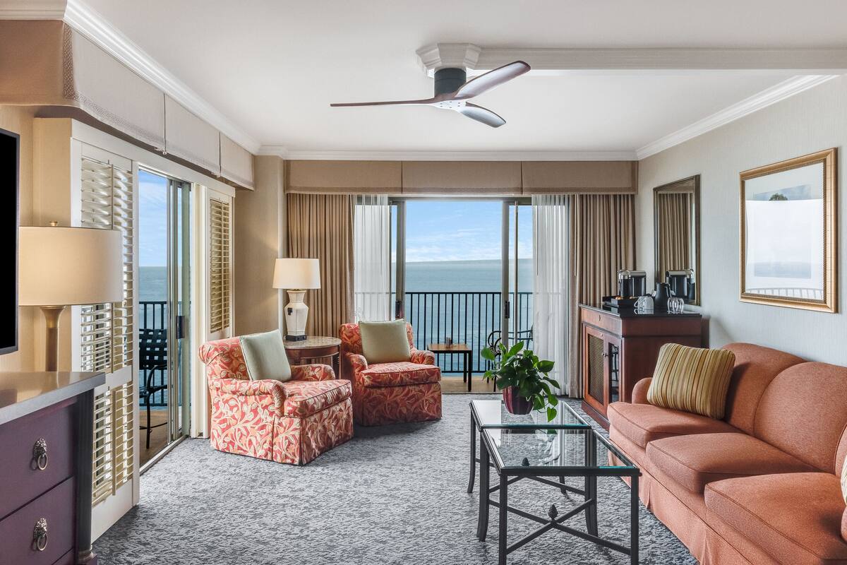 A hotel room with a balcony, a sofa, armchairs, a coffee table, and a television.