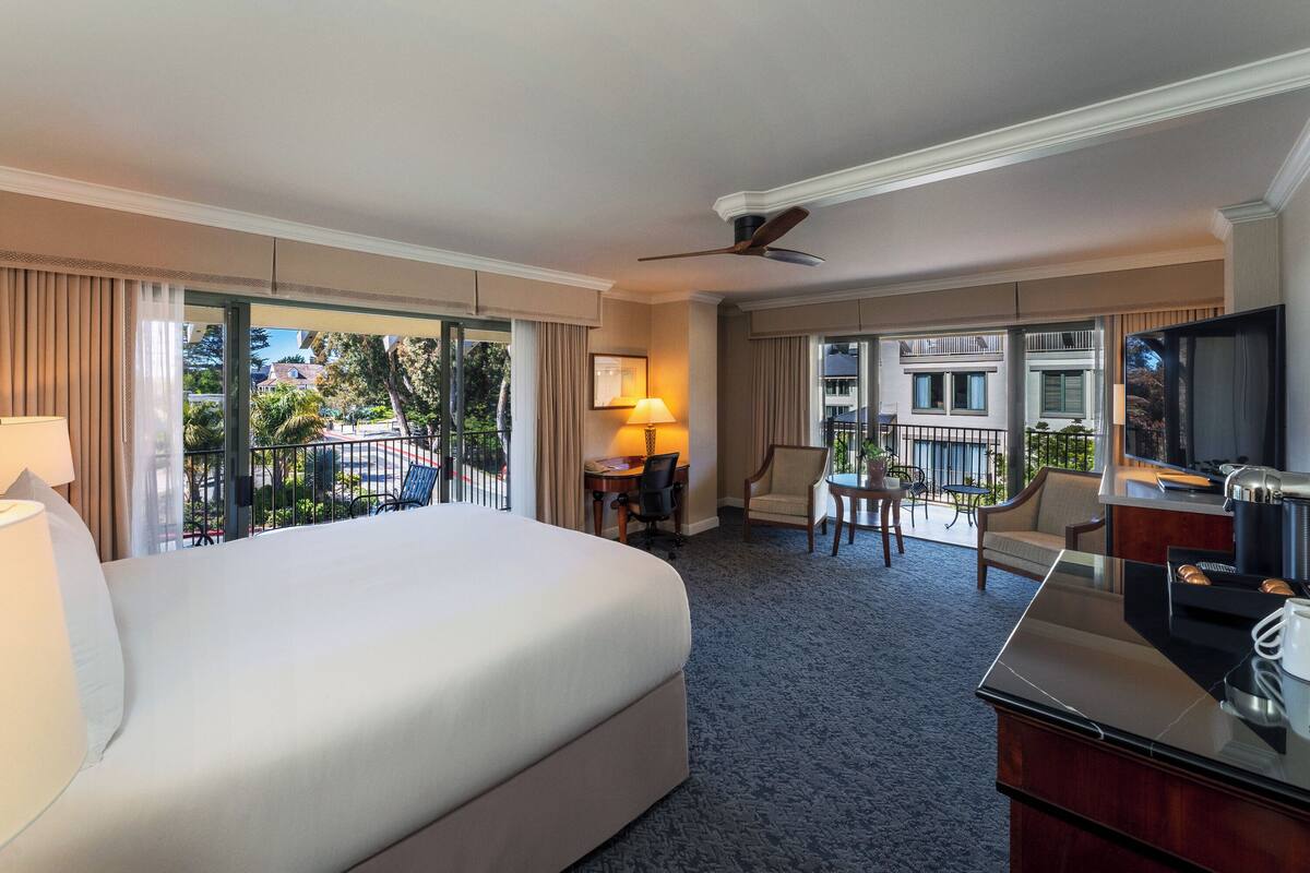 A hotel room with a large bed, a desk with a chair, a television, and a balcony with a view of the city.