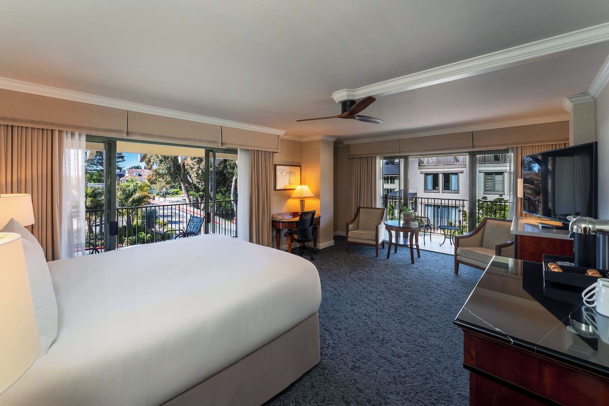 A hotel room with a large bed, a desk with a chair, a television, and a balcony with a view of the city.