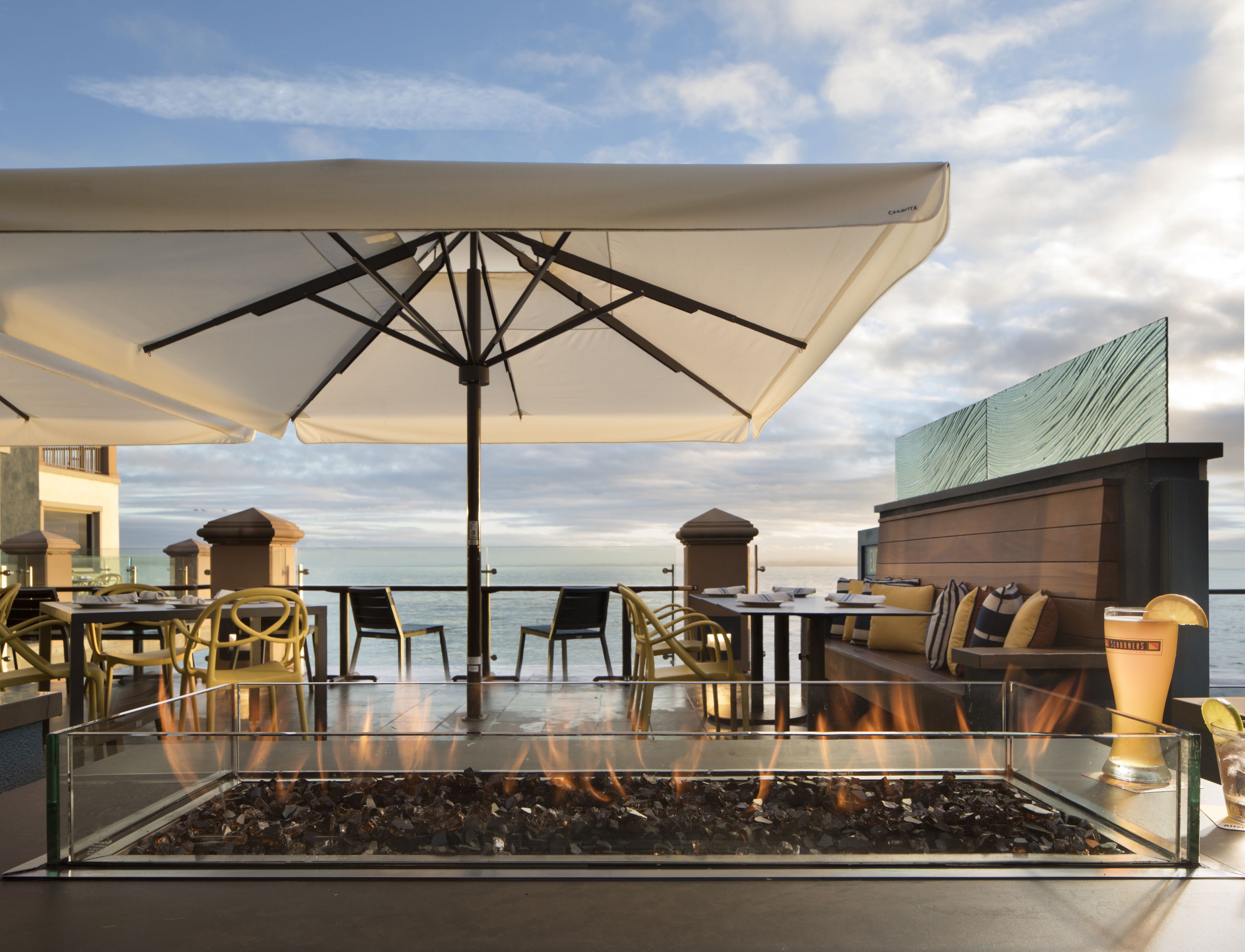 Un espace extérieur aménagé avec des sièges, une table basse surmontée d’un foyer en verre, des tables, des chaises et des parasols.