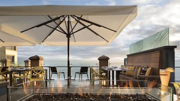 Un espace extérieur aménagé avec des sièges, une table basse surmontée d’un foyer en verre, des tables, des chaises et des parasols.