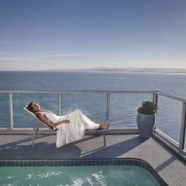 A person relaxing on a balcony with a pool, overlooking a body of water and distant land.