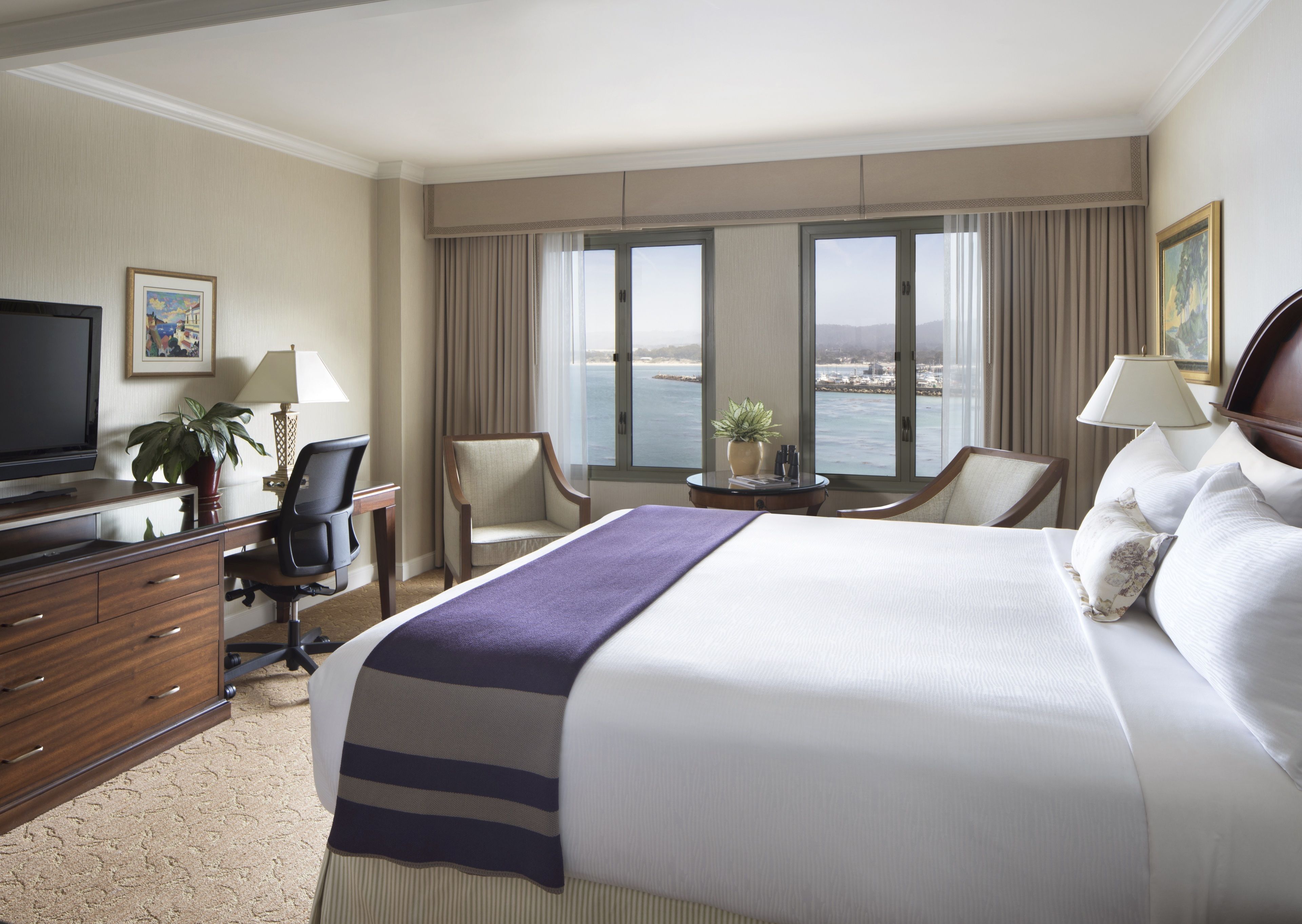 A hotel room with a large bed, a desk with a computer, a chair, a TV, and a view of the sea.