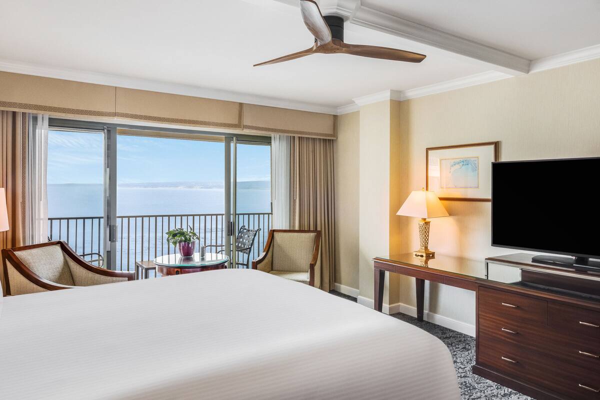 A hotel room with a large bed, a desk with a television, a chair, a small table with a plant, and a view of the sea through large windows.