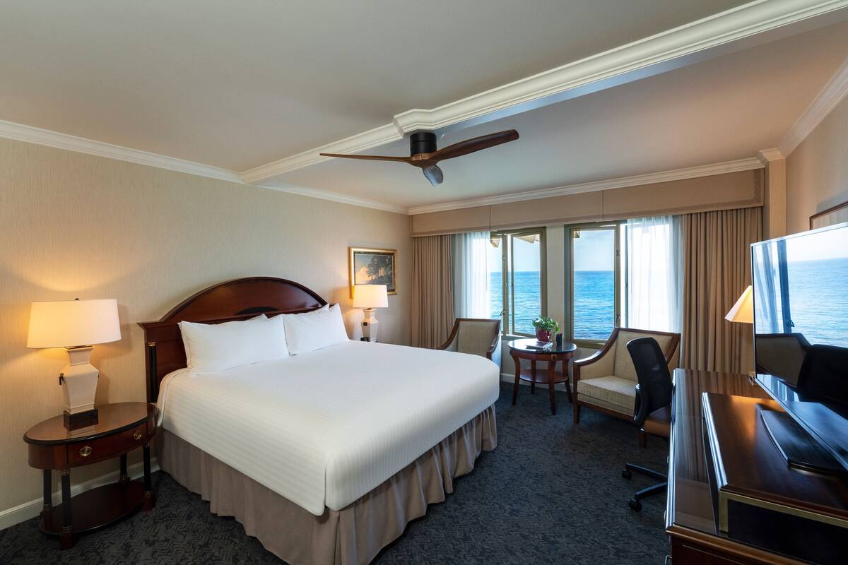 A hotel room with a large bed, a desk, a chair, a television, and a view of the ocean.