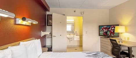 A hotel room with a bed, a desk with a chair, a television, and a bathroom visible through an open door.