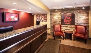 A Red Roof Inn reception area with a granite counter, leather chairs, a small table, and a mounted TV displaying room upgrade information.