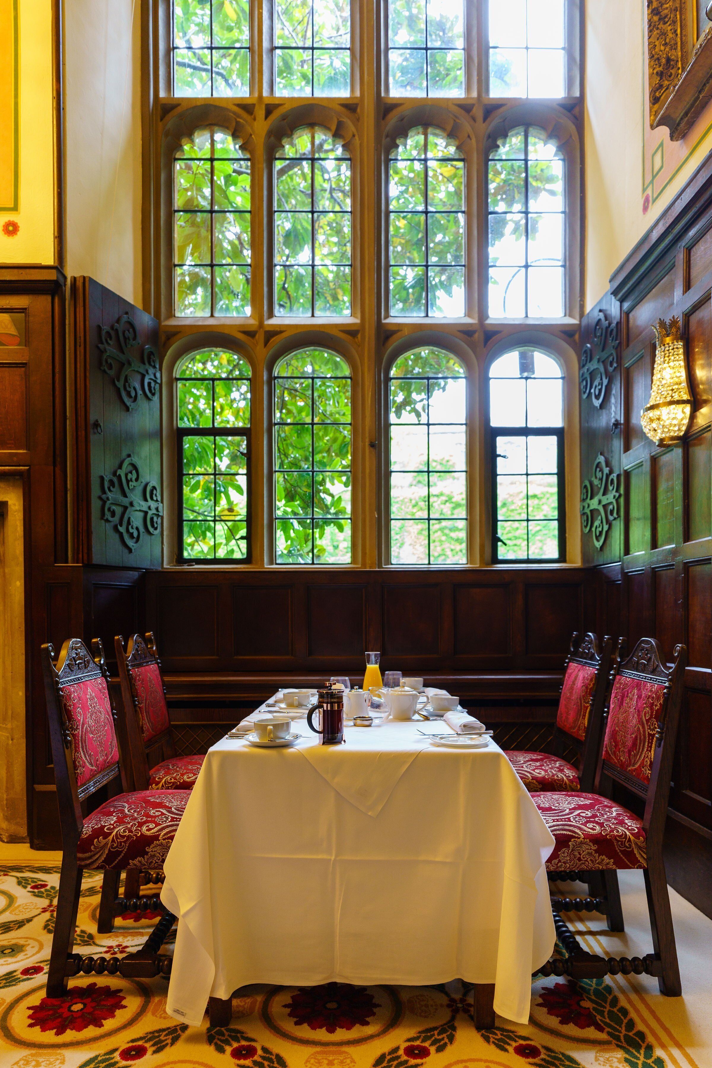 Un coin repas avec une table dressée pour quatre personnes, entouré de chaises décoratives, de grandes fenêtres offrant une vue sur la verdure et de boiseries.