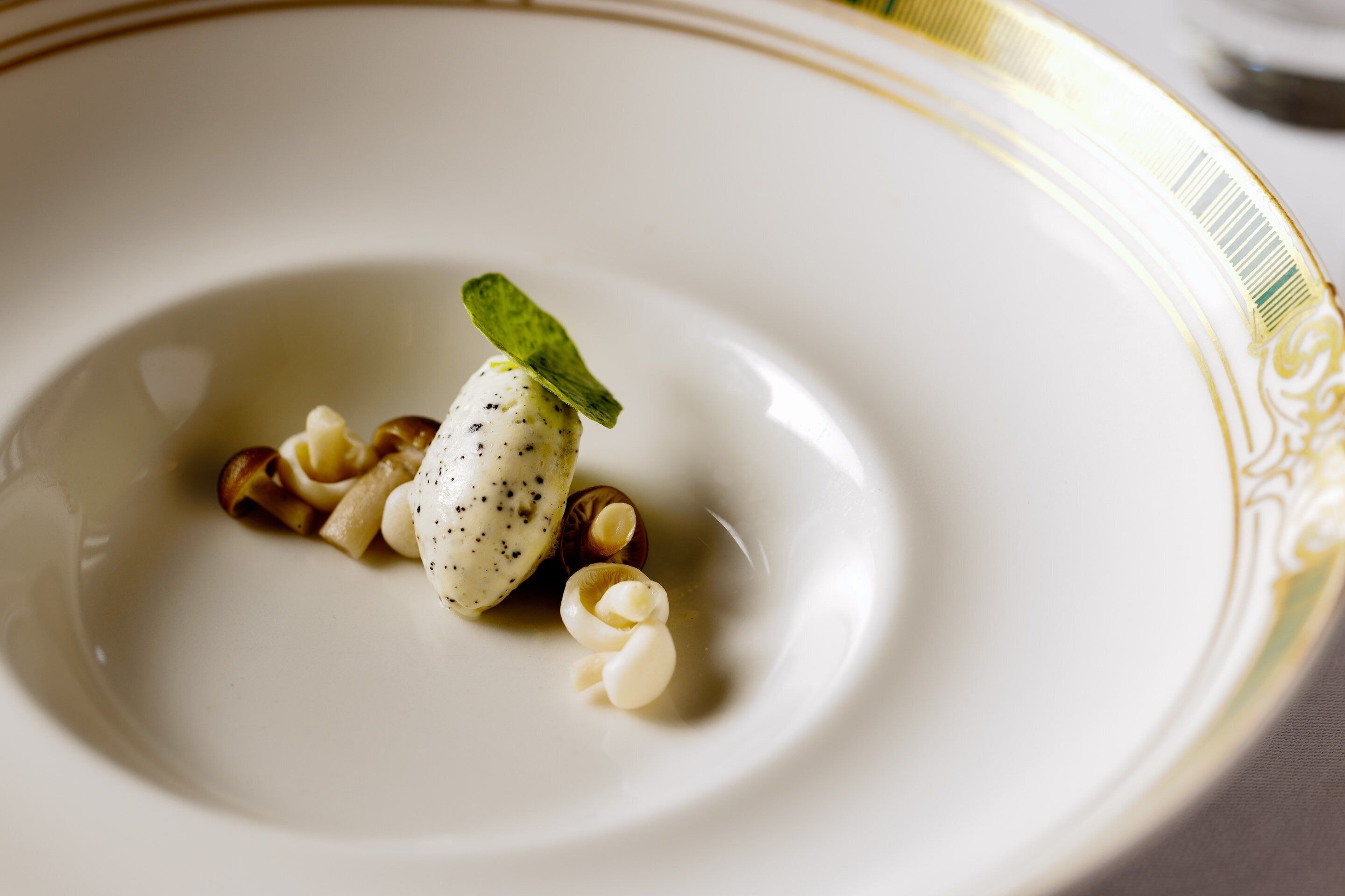 Un plato blanco con un borde decorativo, que contiene un postre con una cucharada de crema y una hoja de menta como adorno.