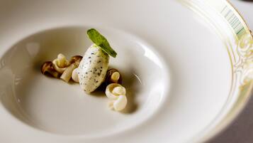 Un plato blanco con un borde decorativo, que contiene un postre con una cucharada de crema y una hoja de menta como adorno.