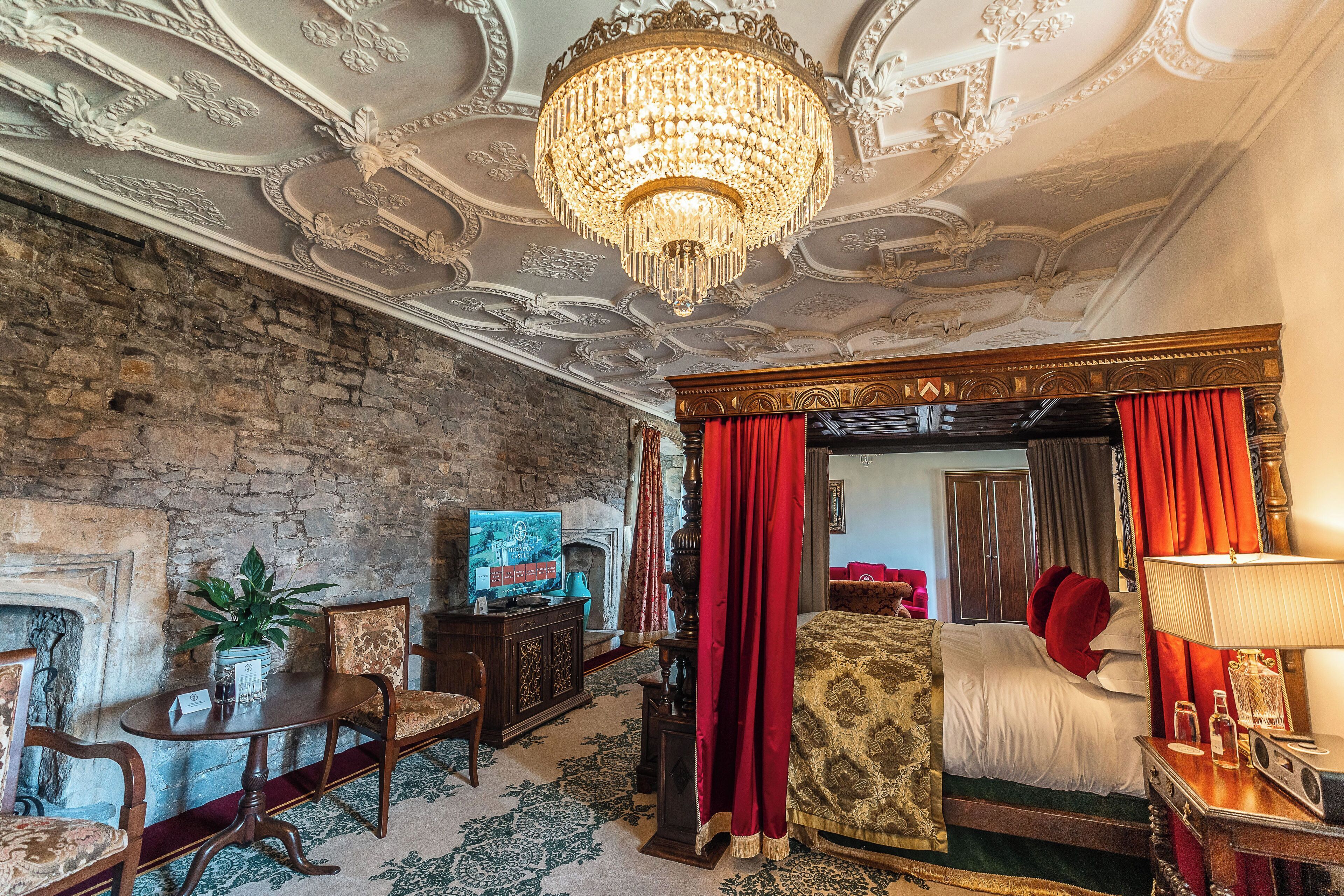 Una habitaci ón con candelabro, chimenea, cama con cortinas rojas y televisor sobre un mueble de madera.