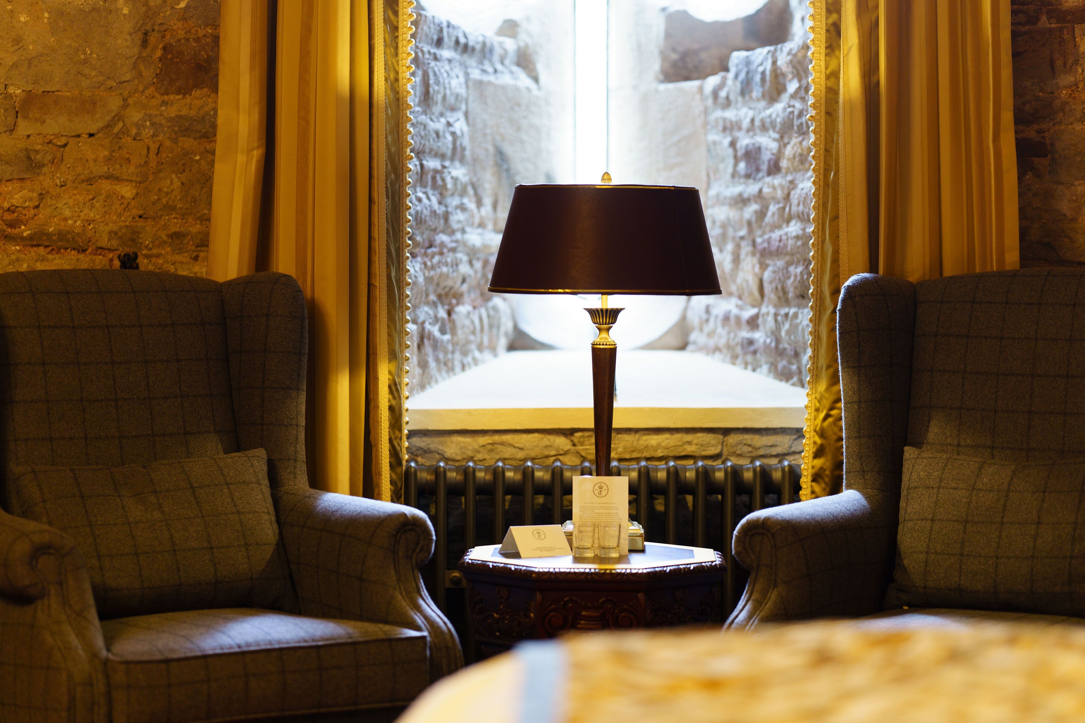 Una habitación con dos sillones, una mesa con lámpara y vistas a un muro de piedra a través de una ventana con cortinas.