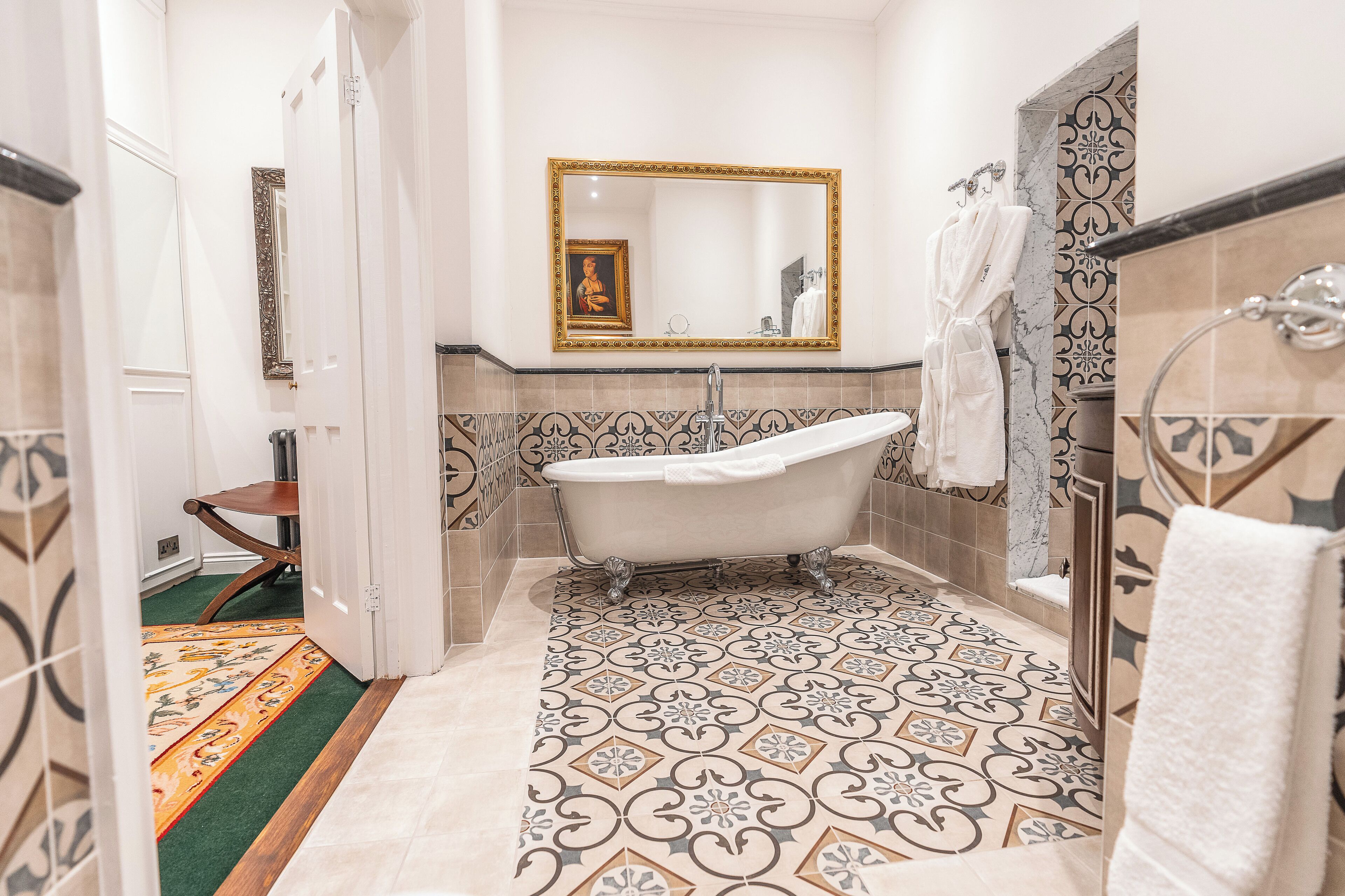 A bathroom with a freestanding bathtub, a large mirror with a decorative frame, and patterned floor tiles.