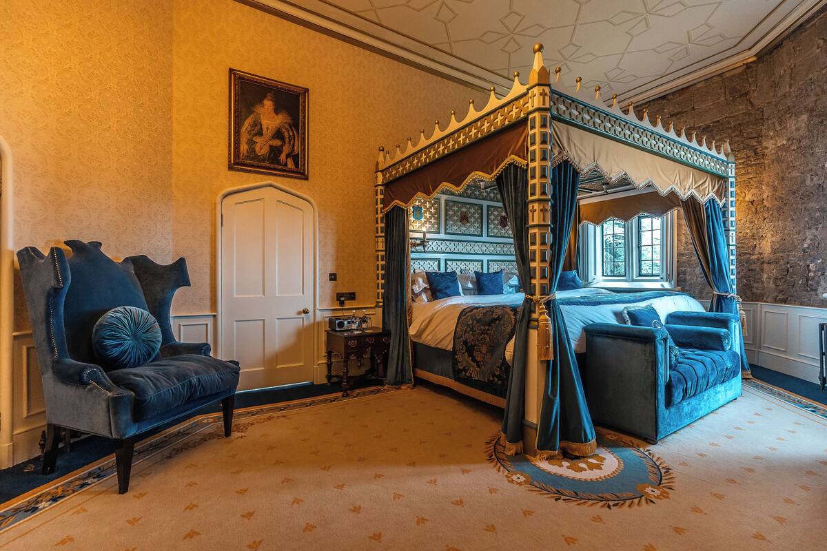 Una cama con dosel, un sillón azul oscuro y una alfombra estampada en una habitación con pared de piedra y una puerta blanca.