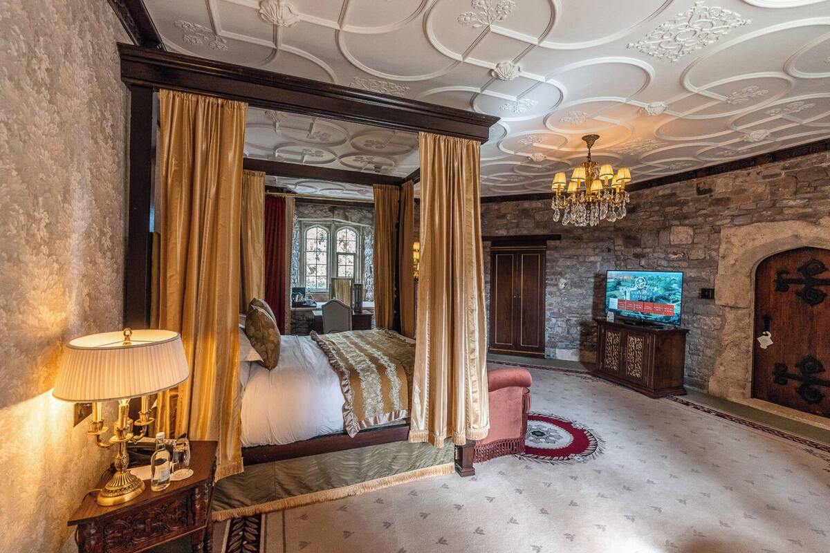 Un dormitorio lujoso con cama con dosel, candelabro, televisor y pared de piedra.