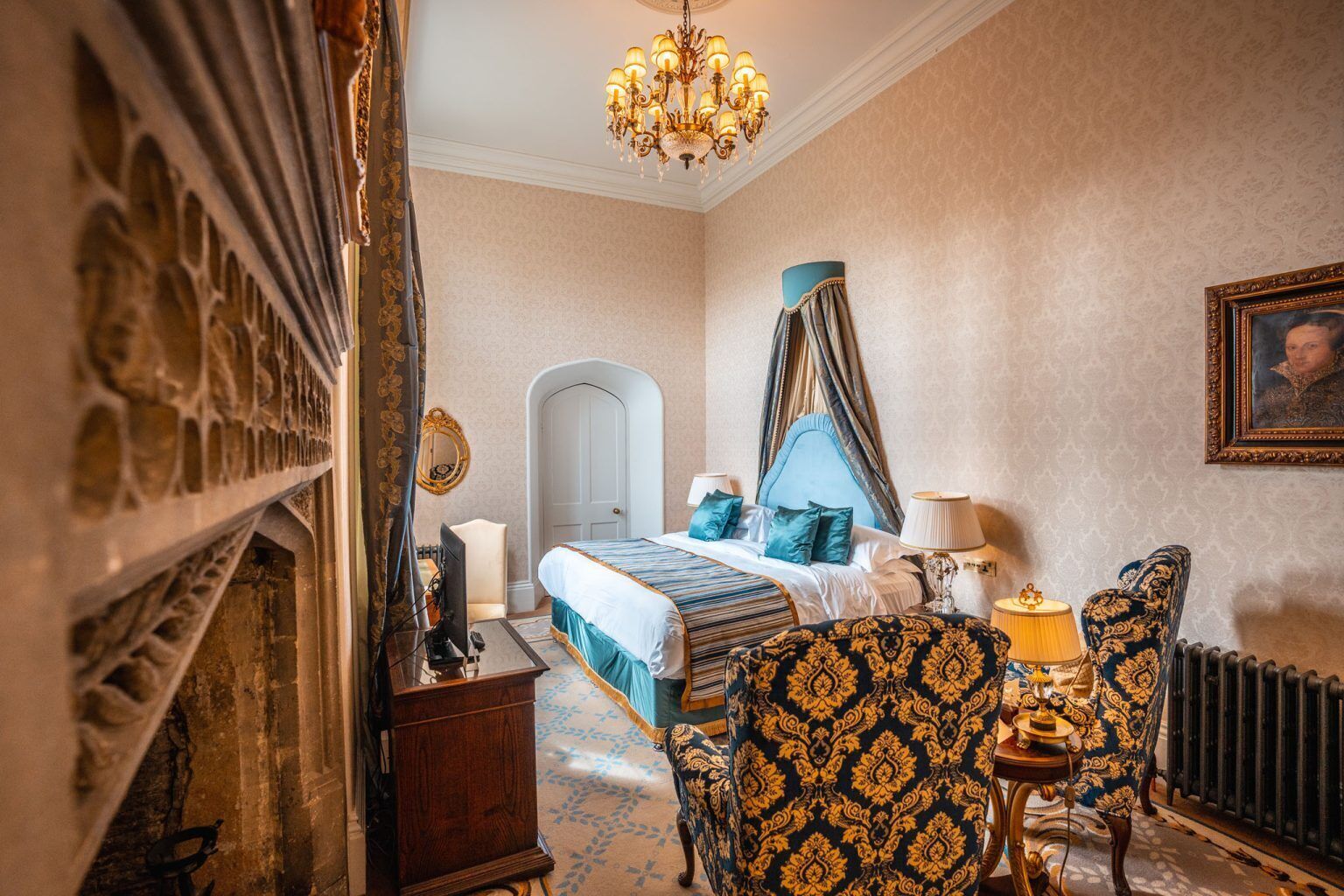 A bedroom with a bed, a desk, a chair, a chandelier, and a framed portrait on the wall.