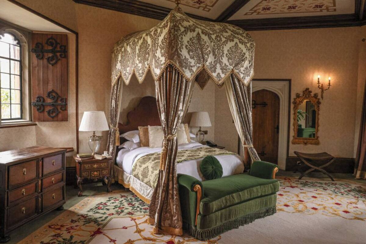 Una cama con dosel y cuatro columnas, un banco tapizado en verde, un aparador de madera y una alfombra con motivos florales.