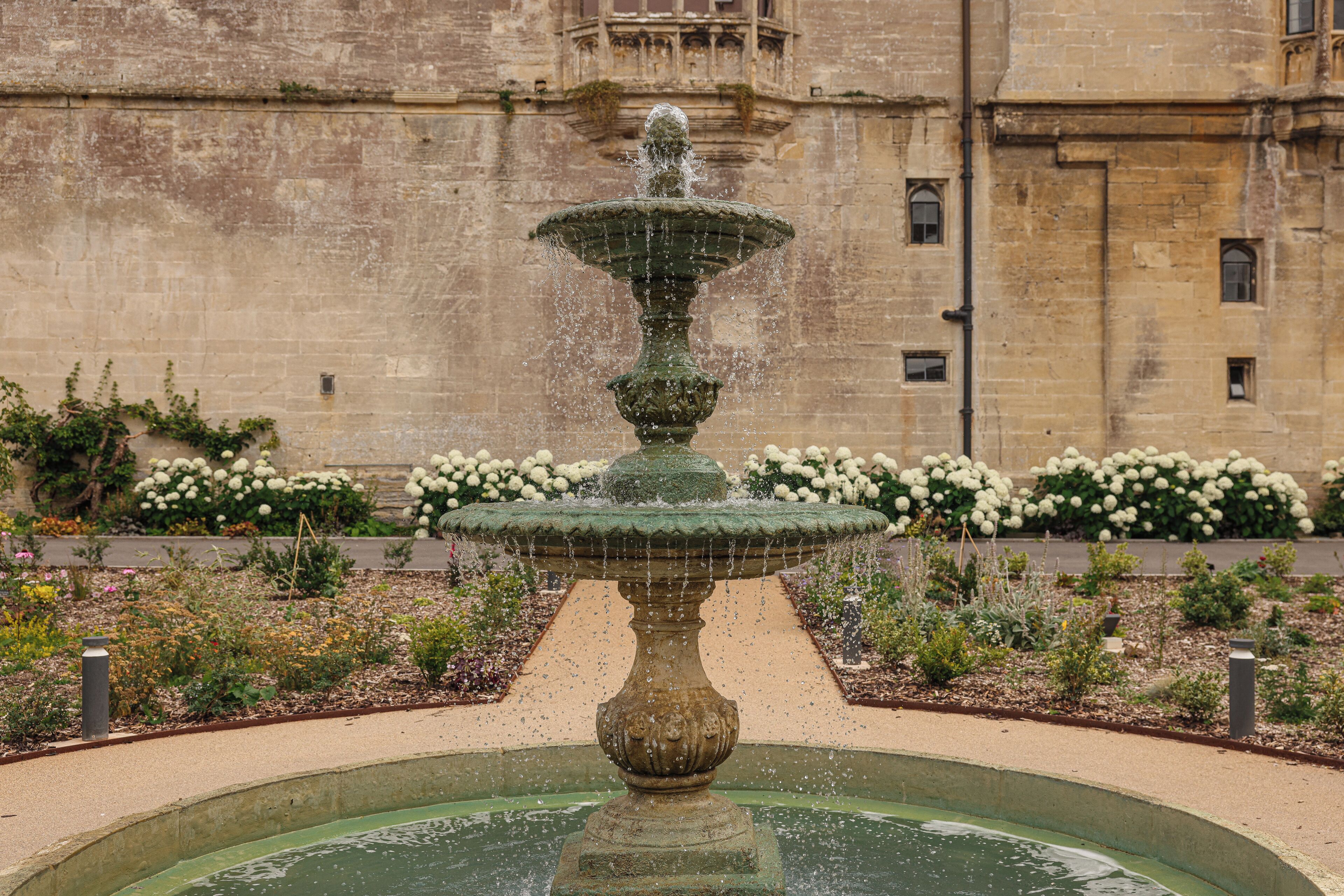 Una fuente de varios niveles en un jardín, con un edificio de piedra al fondo.