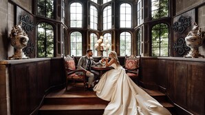 A couple seated in a room with large arched windows, wooden paneling, and ornate vases. - Thornbury Castle, Relais & Châteaux (Bristol)