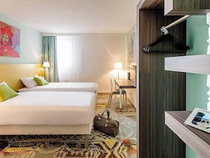 Habitación de hotel con dos camas, un escritorio, una silla, perchero y mesita de noche con lámpara.