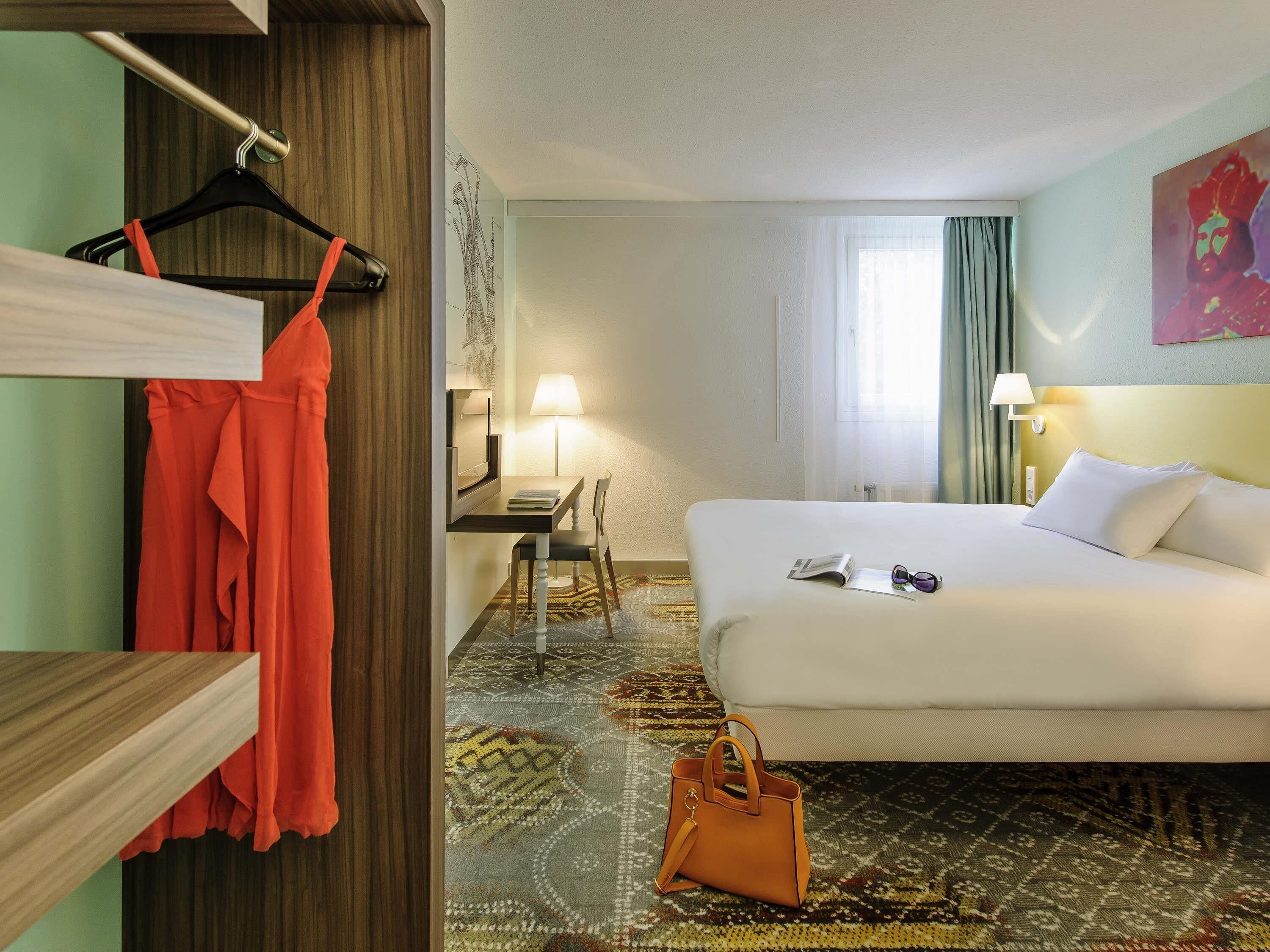 Een hotelkamer met een bed, een bureau, een stoel, een kapstok en een handtas.