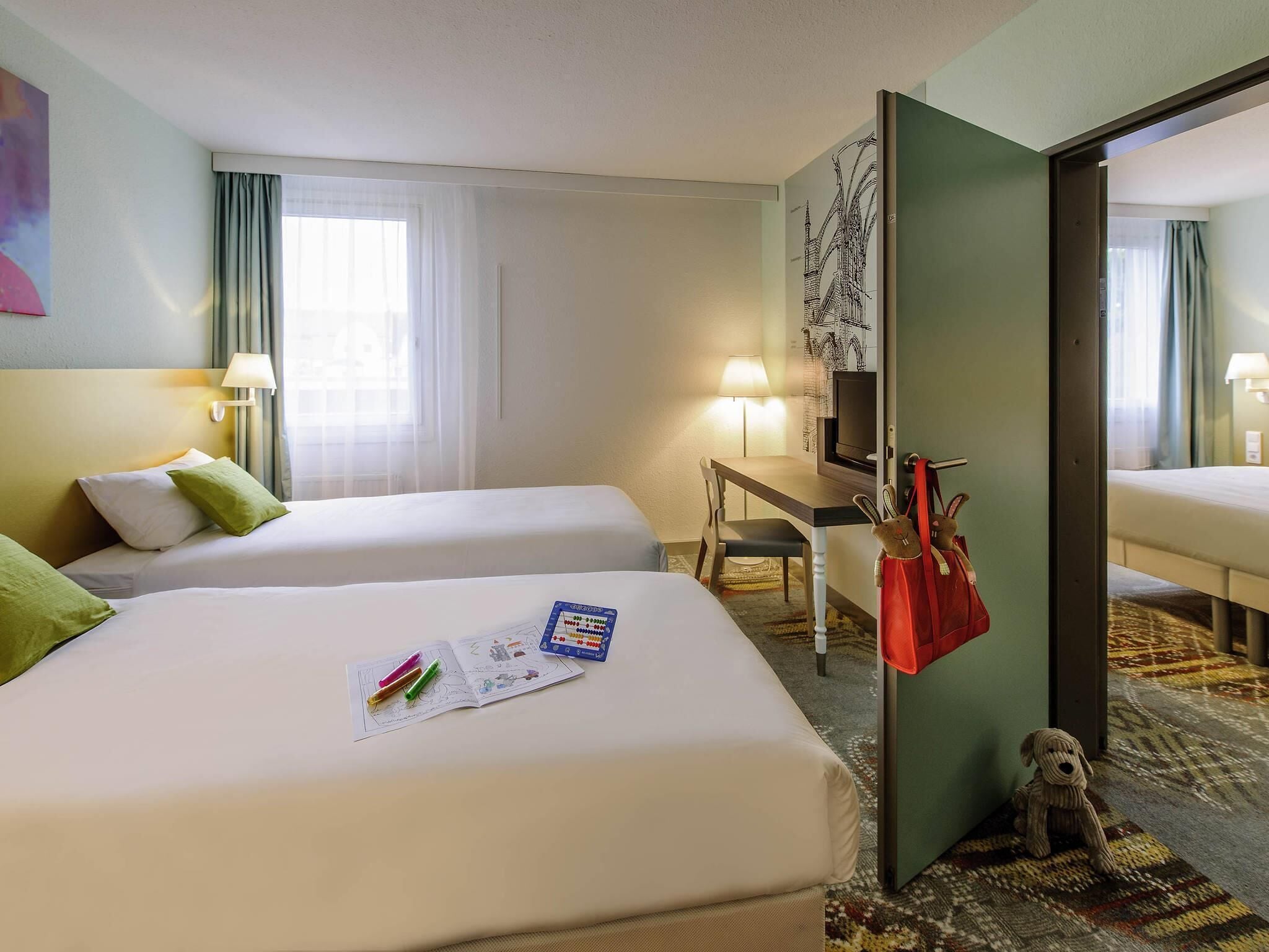 Habitación de hotel con dos camas, un escritorio con silla, televisión y una puerta con un bolso rojo colgando.