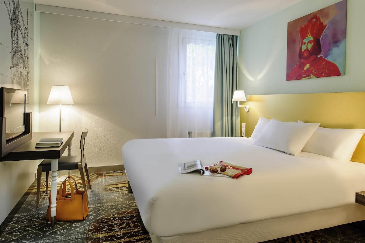Habitación de hotel con cama, escritorio, silla, lámpara, bolso de mano y un cuadro en la pared.