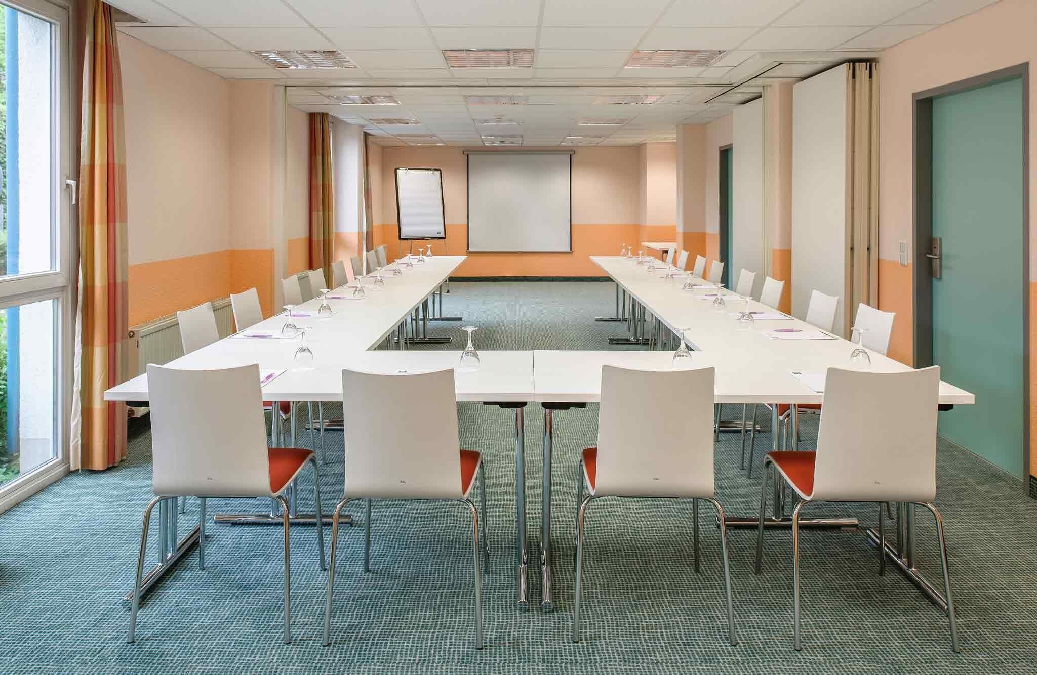 Un salón de conferencias con una larga mesa, sillas blancas y una pantalla para proyector.