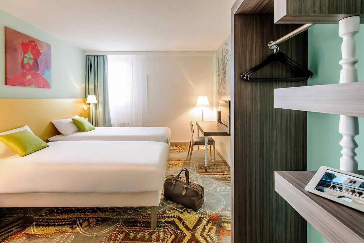 Habitación de hotel con dos camas, un escritorio, una silla, perchero y un marco digital con fotografías en un estante.