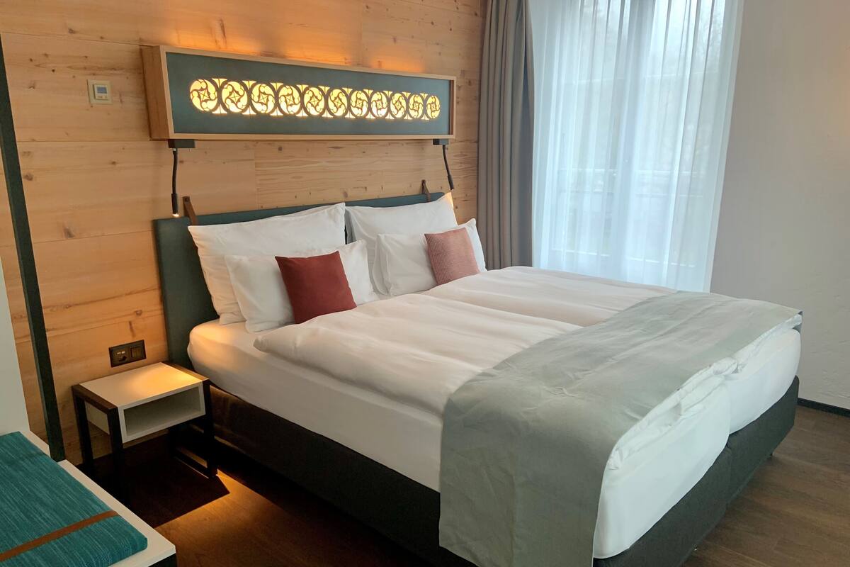 A neatly made double bed with white and red pillows, a bedside table with a lamp, and a decorative wall panel above the bed.