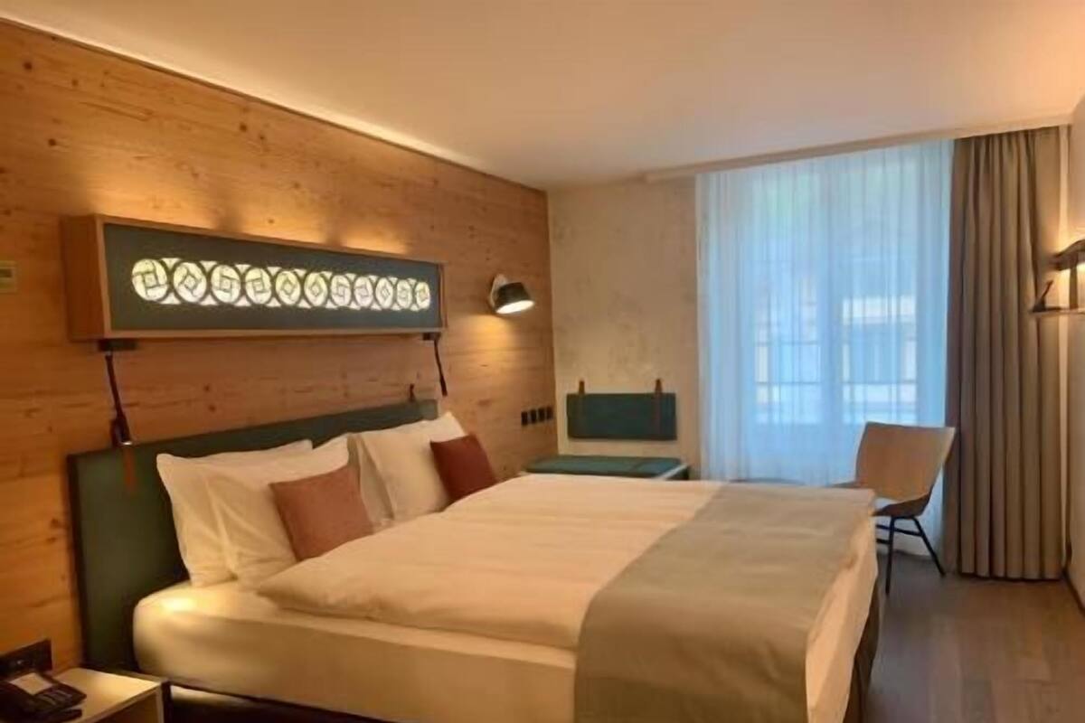 A hotel room with a bed, bedside table, lamp, chair, and a window with curtains.