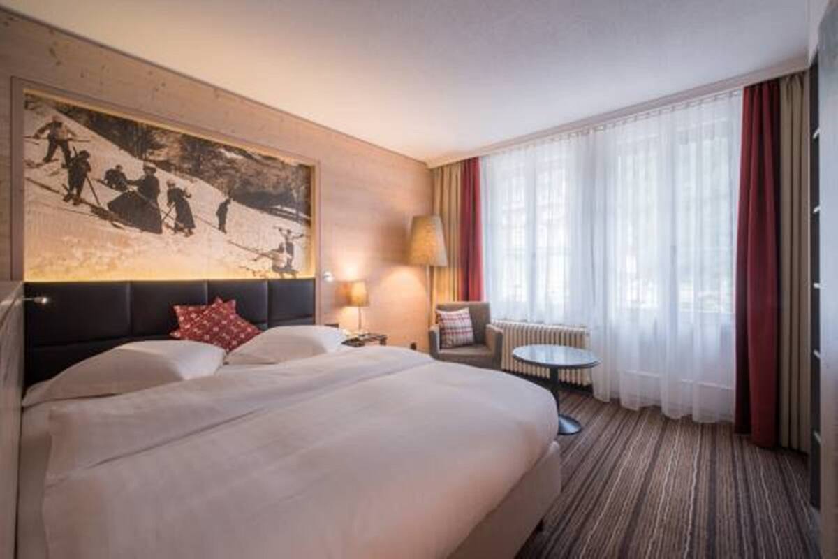A hotel room with a large bed, a painting on the wall, a small table, a chair, and a window with curtains.